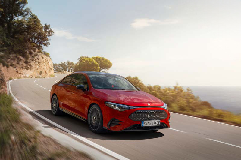 Eine rote Mercedes-Benz-Limousine fährt auf einer kurvenreichen Küstenstraße mit Bäumen und Klippen auf der einen Seite und dem Meer im Hintergrund unter einem klaren Himmel.