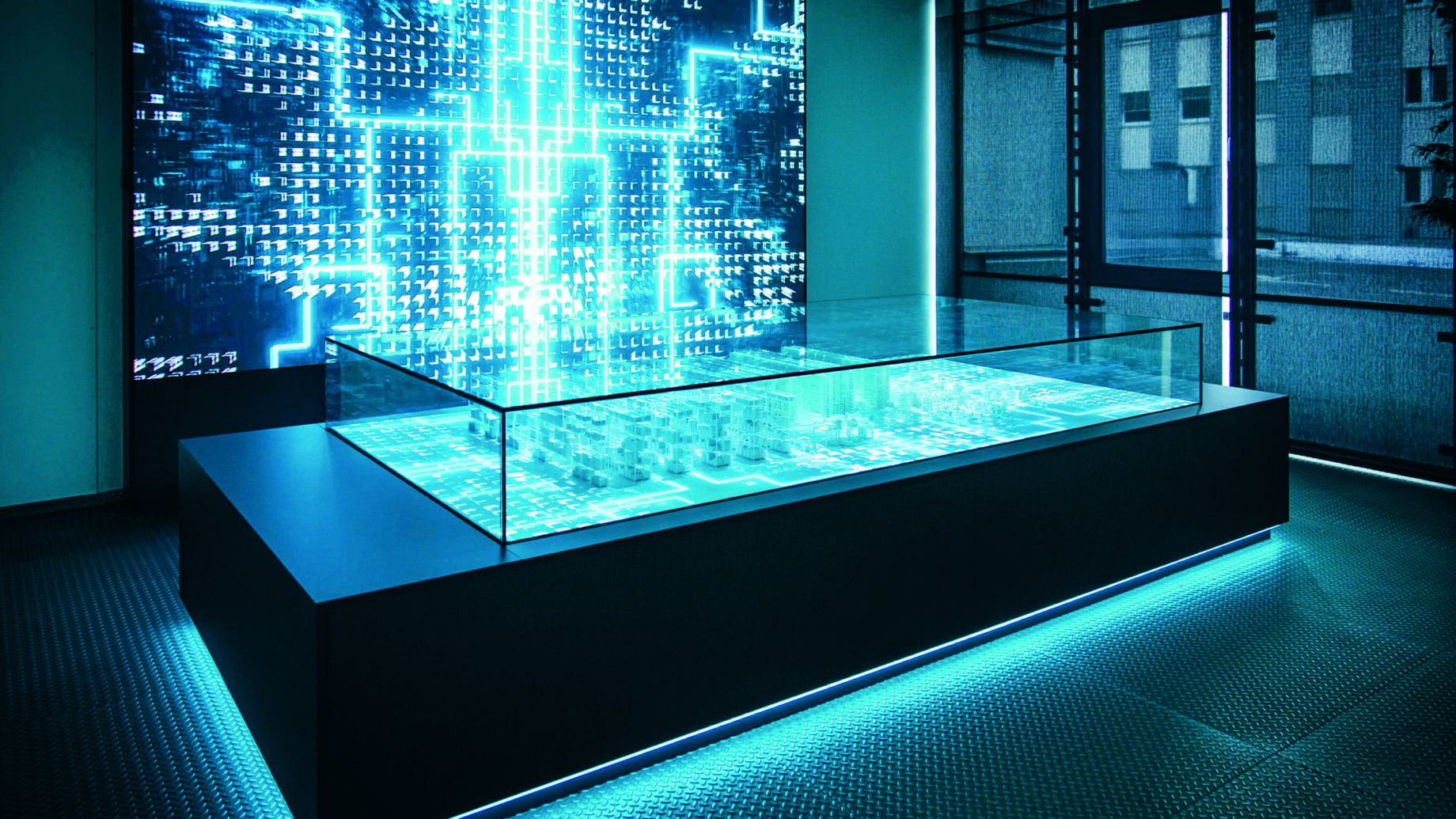 In einem modernen Hightech-Raum steht ein großer beleuchteter Tisch mit einer Glasplatte, auf der ein digitales Modell zu sehen ist, vor einer leuchtenden Wand mit blauen Schaltkreisen und Zahlen, die eine futuristische Atmosphäre schaffen.