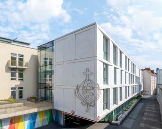 Ein modernes weißes Gebäude mit großen Fenstern und einem gläsernen Eingang steht neben einem hellbraunen Gebäude; an der Fassade befindet sich ein dekoratives Emblem, und an der unteren Wand sind bunte geometrische Muster aufgemalt.