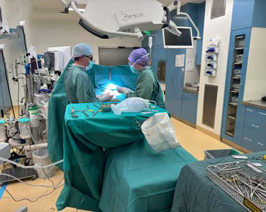 Zwei Chirurgen in Kitteln und Masken führen eine Operation in einem Operationssaal durch, der mit medizinischen Geräten, chirurgischen Werkzeugen und hellen Deckenleuchten ausgestattet ist. Ein Patient liegt auf dem Tisch, der mit grünen OP-Tüchern abgedeckt ist.
