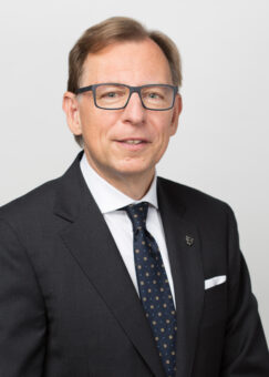 Ein Mann mittleren Alters mit kurzen hellbraunen Haaren und Brille, der einen dunklen Anzug, ein weißes Hemd und eine gemusterte marineblaue Krawatte trägt, posiert vor einem schlichten hellen Hintergrund.