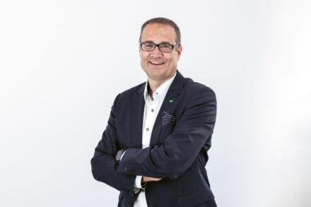 Ein Mann, der eine Brille, einen dunklen Blazer und ein weißes Hemd trägt, steht vor einem schlichten weißen Hintergrund und lächelt mit verschränkten Armen.