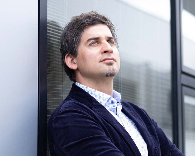 Ein Mann mit kurzen dunklen Haaren und einem Spitzbart, der einen marineblauen Blazer und ein gemustertes Hemd trägt, lehnt an einer Glaswand und blickt nachdenklich nach oben.