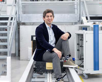 Ein Mann in einem dunklen Blazer, einem gemusterten Hemd, einer grauen Hose und bunten Socken sitzt an einer Industrieausrüstung in einem hellen, modernen Labor mit Metalltreppen und Maschinen im Hintergrund.