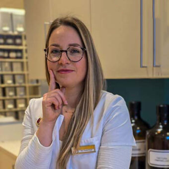 Eine Frau mit langen blonden Haaren und Brille, die einen weißen Kittel und ein Namensschild trägt, steht mit einem Finger am Kinn in einem Raum und lächelt leicht. Im Hintergrund sind große braune Flaschen und Regale zu sehen.