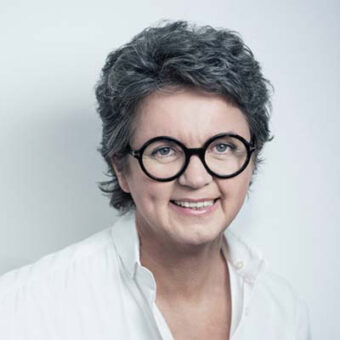 Eine lächelnde Person mit kurzen, lockigen grauen Haaren und einer runden schwarzen Brille, die ein weißes Hemd mit Kragen trägt, posiert vor einem schlichten hellen Hintergrund.