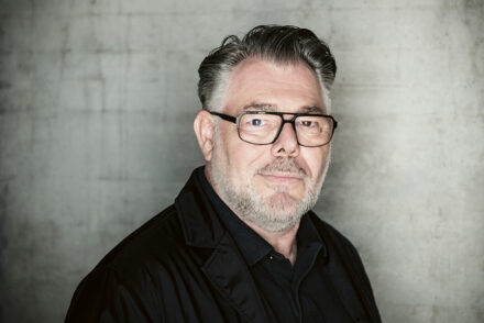 Ein Mann mittleren Alters mit grauem Haar und Bart, der eine schwarze Brille, ein dunkles Hemd mit Kragen und eine schwarze Jacke trägt, steht vor einer schlichten grauen, strukturierten Wand und blickt ruhig in die Kamera.