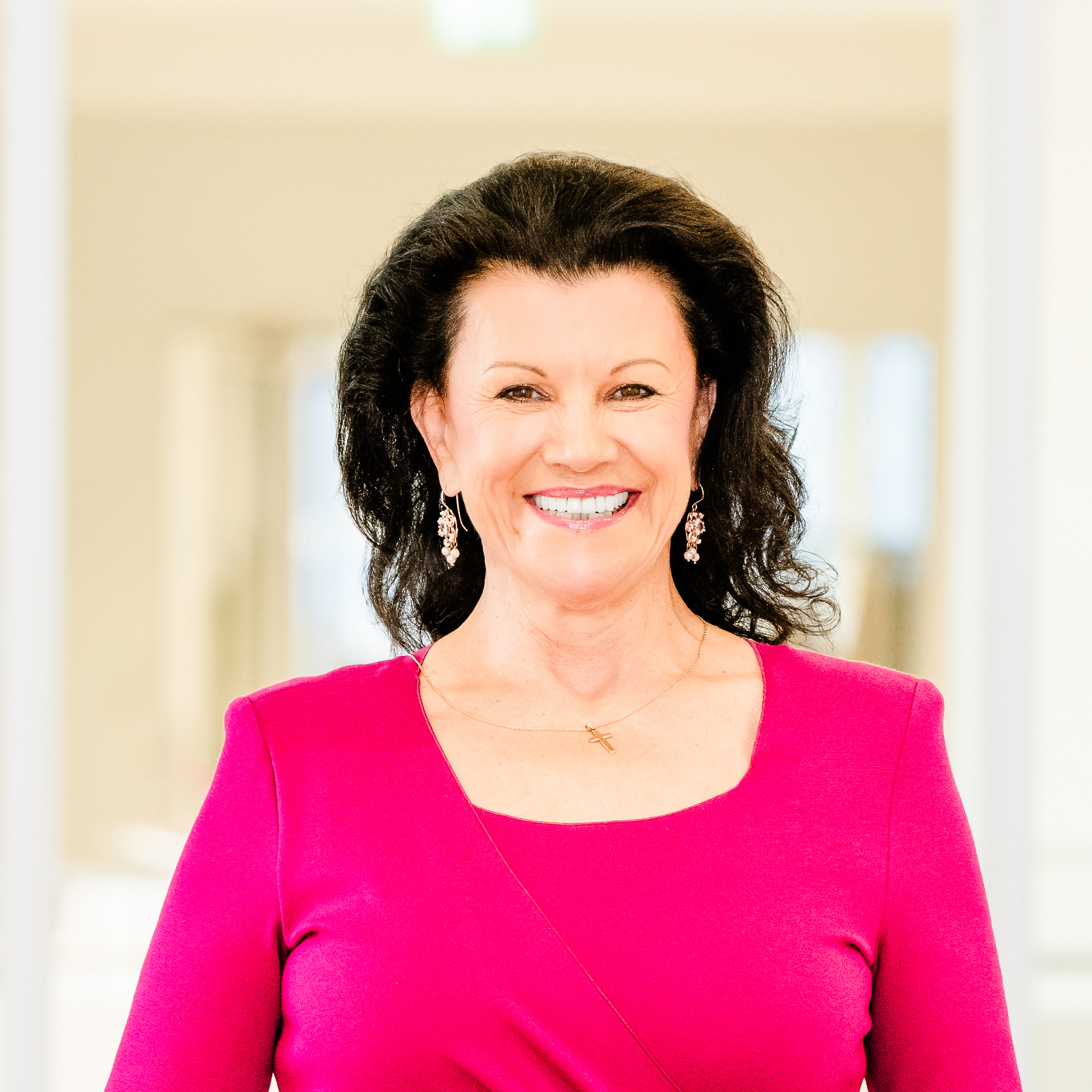 Eine Frau mit dunklem, gewelltem Haar lächelt und trägt ein hellrosa Kleid und baumelnde Ohrringe. Sie steht in einem sanft beleuchteten Innenraum mit unscharfem Hintergrund.