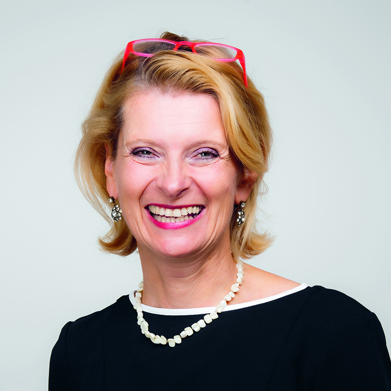 Lächelnde Frau mit kurzen blonden Haaren, die ein schwarzes Oberteil mit weißem Saum, eine Perlenkette, Ohrringe und eine rote Brille auf dem Kopf trägt, posiert vor einem schlichten hellen Hintergrund.