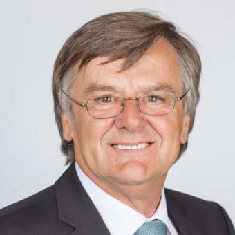 Ein Mann mittleren Alters mit grauem Haar und Brille, der einen dunklen Anzug, ein weißes Hemd und eine hellblaue Krawatte trägt, lächelt in die Kamera vor einem schlichten hellen Hintergrund.