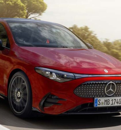 Eine rote Mercedes-Benz-Limousine fährt an einem sonnigen Tag eine kurvenreiche Küstenstraße entlang, während im Hintergrund das Meer und die Bäume zu sehen sind.
