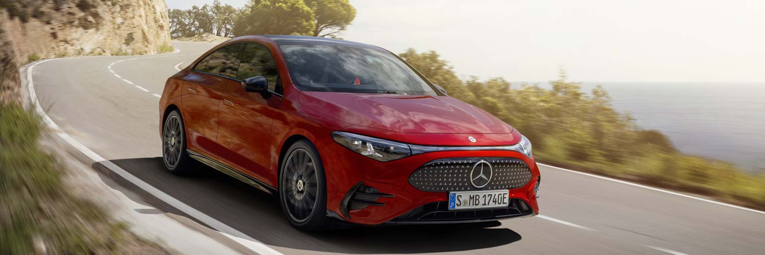 Eine rote Mercedes-Benz-Limousine fährt an einem sonnigen Tag eine kurvenreiche Küstenstraße entlang, während im Hintergrund das Meer und die Bäume zu sehen sind.