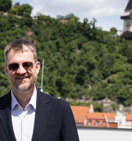 Ein Mann in einem dunklen Anzug und mit Sonnenbrille steht lächelnd im Freien, mit roten Dächern und einem üppig grünen Hügel mit einem Uhrenturm im Hintergrund unter einem teilweise bewölkten Himmel.
