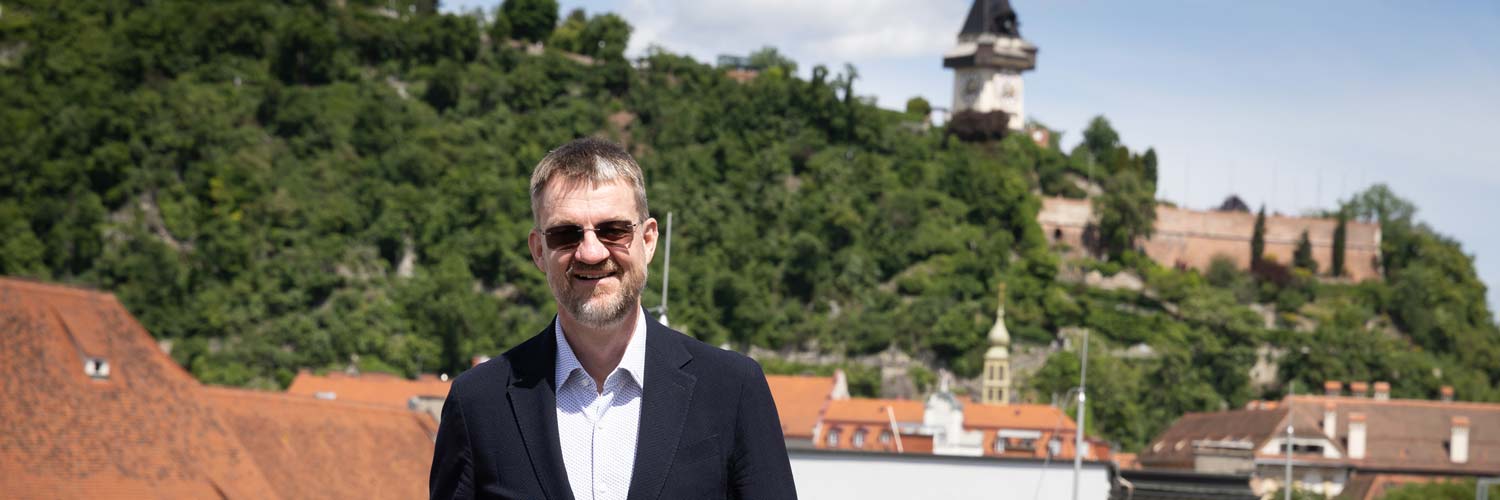 Ein Mann in einem dunklen Anzug und mit Sonnenbrille steht lächelnd im Freien, mit roten Dächern und einem üppig grünen Hügel mit einem Uhrenturm im Hintergrund unter einem teilweise bewölkten Himmel.