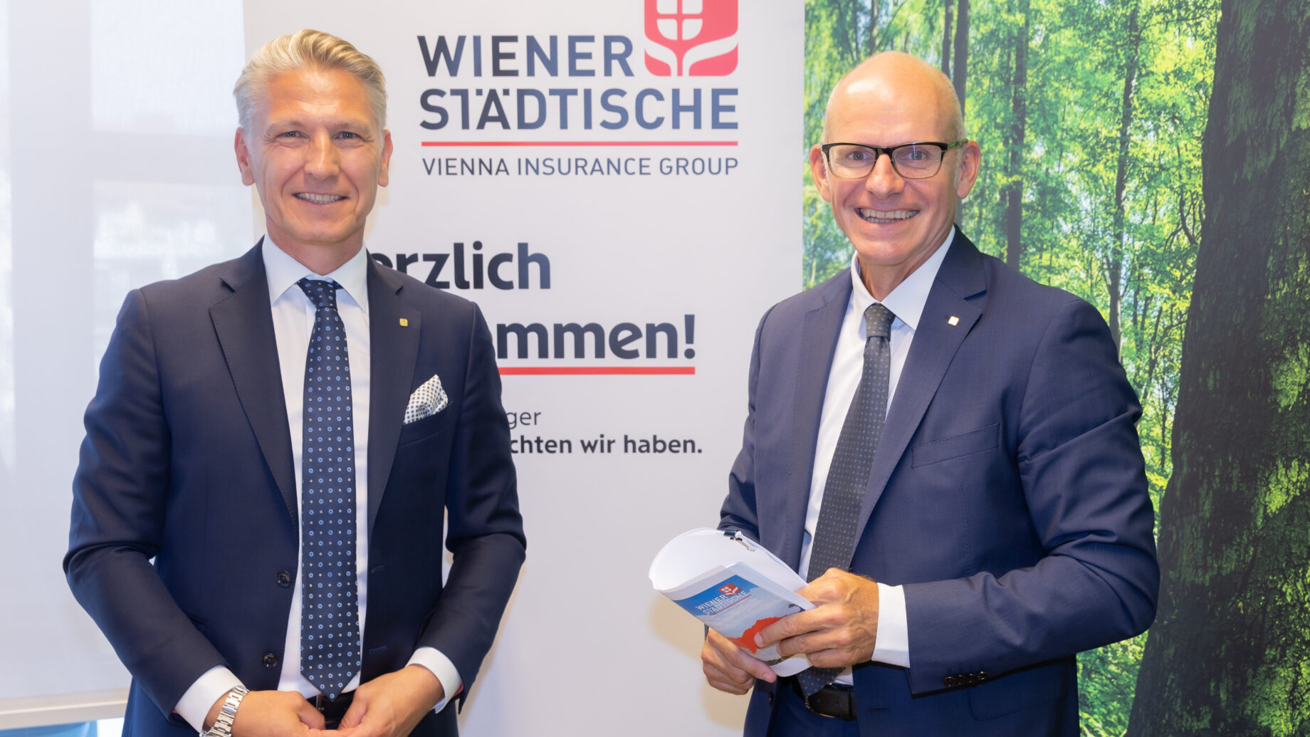 Zwei Männer in Geschäftsanzügen stehen lächelnd vor einem Versicherungsbanner der Wiener Städtischen und einer Waldwand, der eine hält Urkunden - Symbol für nachhaltiges Wachstum und Partnerschaft.