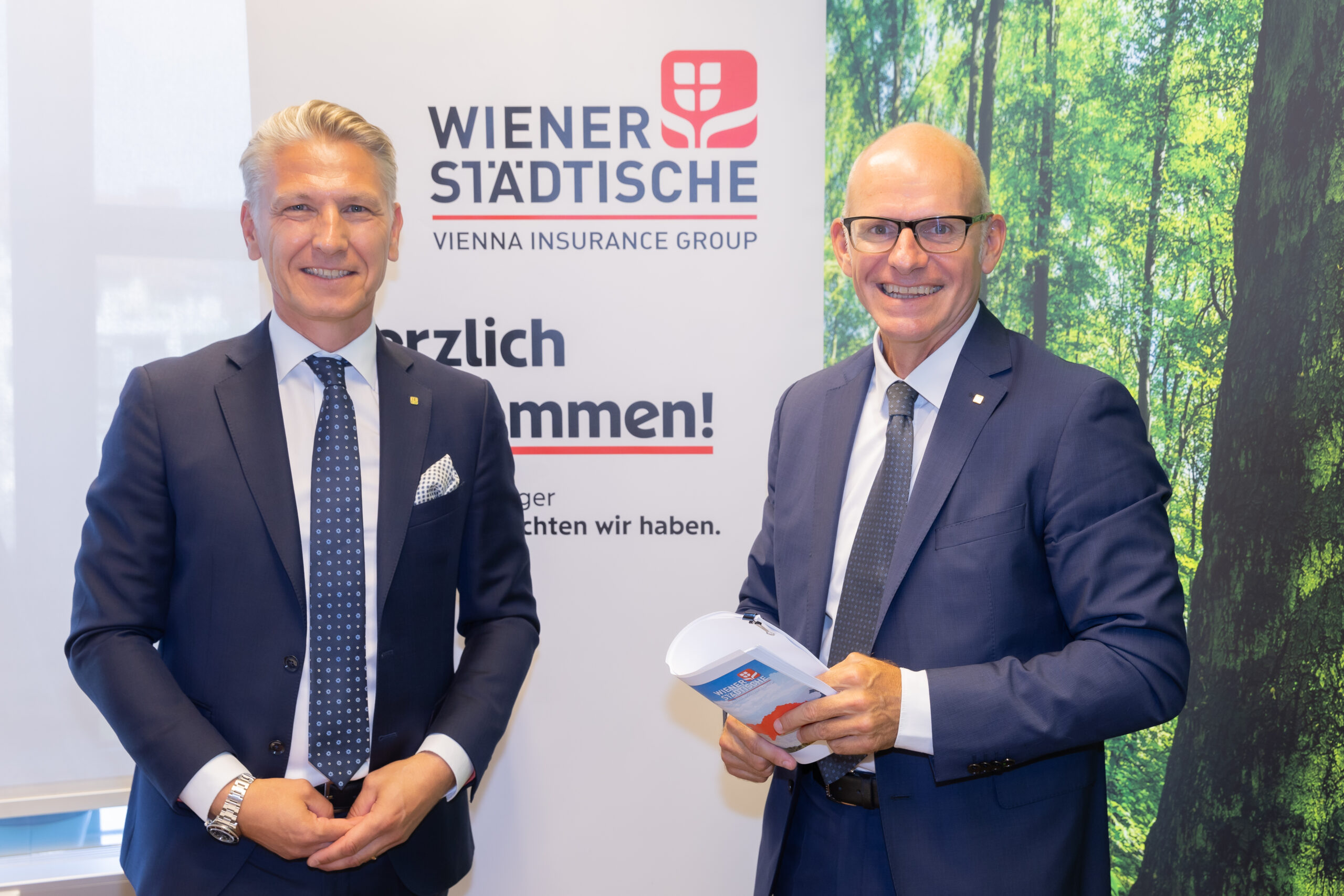 Zwei Männer in Geschäftsanzügen stehen lächelnd vor einem Versicherungsbanner der Wiener Städtischen und einer Waldwand, der eine hält Urkunden - Symbol für nachhaltiges Wachstum und Partnerschaft.