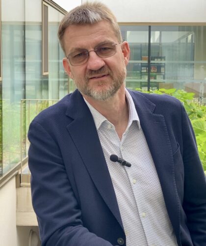 Ein Mann mit kurzen Haaren, Brille und Bart, der einen dunklen Blazer, ein helles Hemd und ein Ansteckmikrofon trägt, steht in einem modernen Gebäudekorridor neben Grünpflanzen und großen Fenstern.