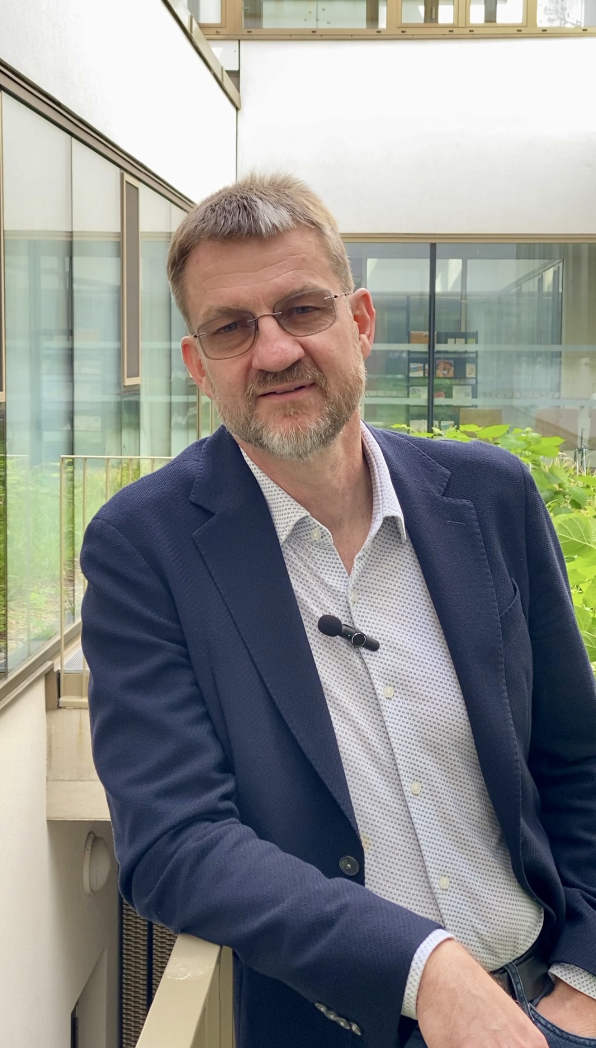 Ein Mann mit kurzen Haaren, Brille und Bart, der einen dunklen Blazer, ein helles Hemd und ein Ansteckmikrofon trägt, steht in einem modernen Gebäudekorridor neben Grünpflanzen und großen Fenstern.