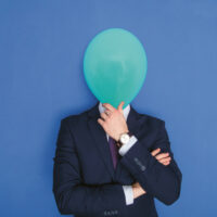 Eine Person in einem marineblauen Anzug steht vor einem blauen Hintergrund und hält sich einen grünen Luftballon vor das Gesicht, um ihre Identität zu verbergen. Ihre Arme sind verschränkt, und sie trägt eine Uhr am linken Handgelenk.