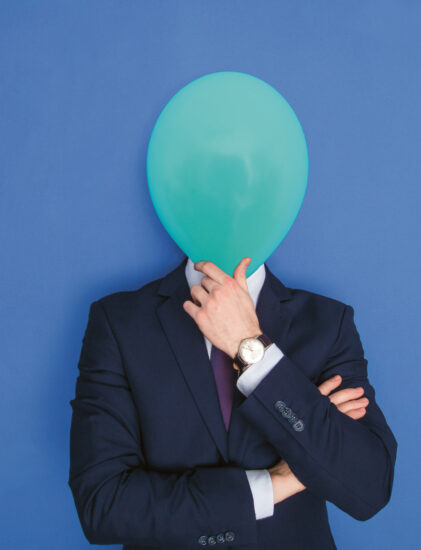 Eine Person in einem marineblauen Anzug steht vor einem blauen Hintergrund und hält sich einen grünen Luftballon vor das Gesicht, um ihre Identität zu verbergen. Ihre Arme sind verschränkt, und sie trägt eine Uhr am linken Handgelenk.