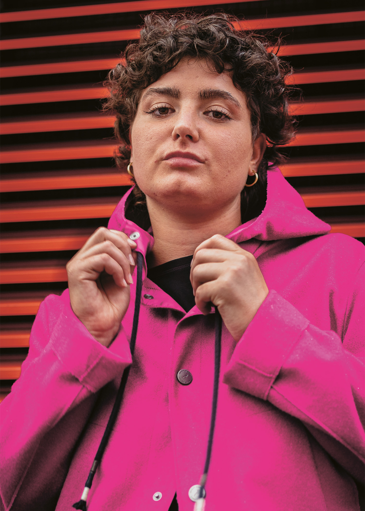 Eine Person mit kurzen lockigen Haaren trägt eine hellrosa Kapuzenjacke, hält den Kragen mit beiden Händen und steht vor einem Hintergrund mit roten und schwarzen horizontalen Lamellen.