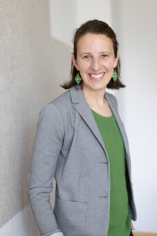 Eine Frau mit braunen Haaren, die einen grauen Blazer über einem grünen Oberteil und grüne geometrische Ohrringe trägt, steht in einem Haus und lächelt in die Kamera.