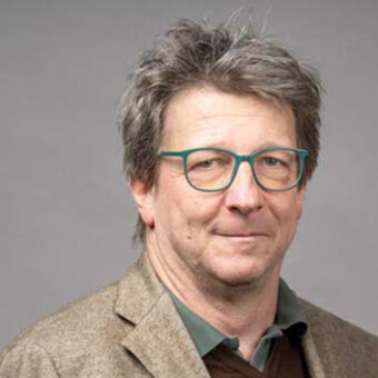 Ein Mann mittleren Alters mit zerzaustem grauem Haar, der eine blaugrüne Brille, einen braunen Blazer und ein grünes Hemd mit Kragen trägt, posiert vor einem schlichten grauen Hintergrund.