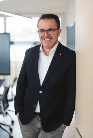 Ein Mann in einem dunklen Blazer, weißem Hemd und Brille lehnt lächelnd an einer Wand in einem modernen Büro mit großen Fenstern und Konferenzraummöbeln.