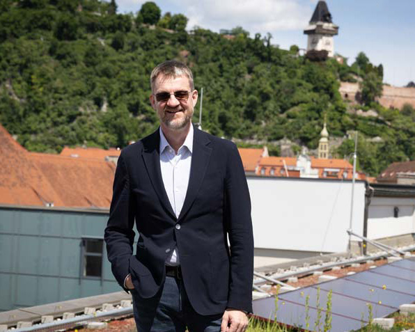 Ein Mann in einem dunklen Blazer und mit Sonnenbrille steht im Freien und lächelt. Im Hintergrund sind eine hügelige, grüne Landschaft und historische Gebäude, darunter ein Uhrenturm, zu sehen.