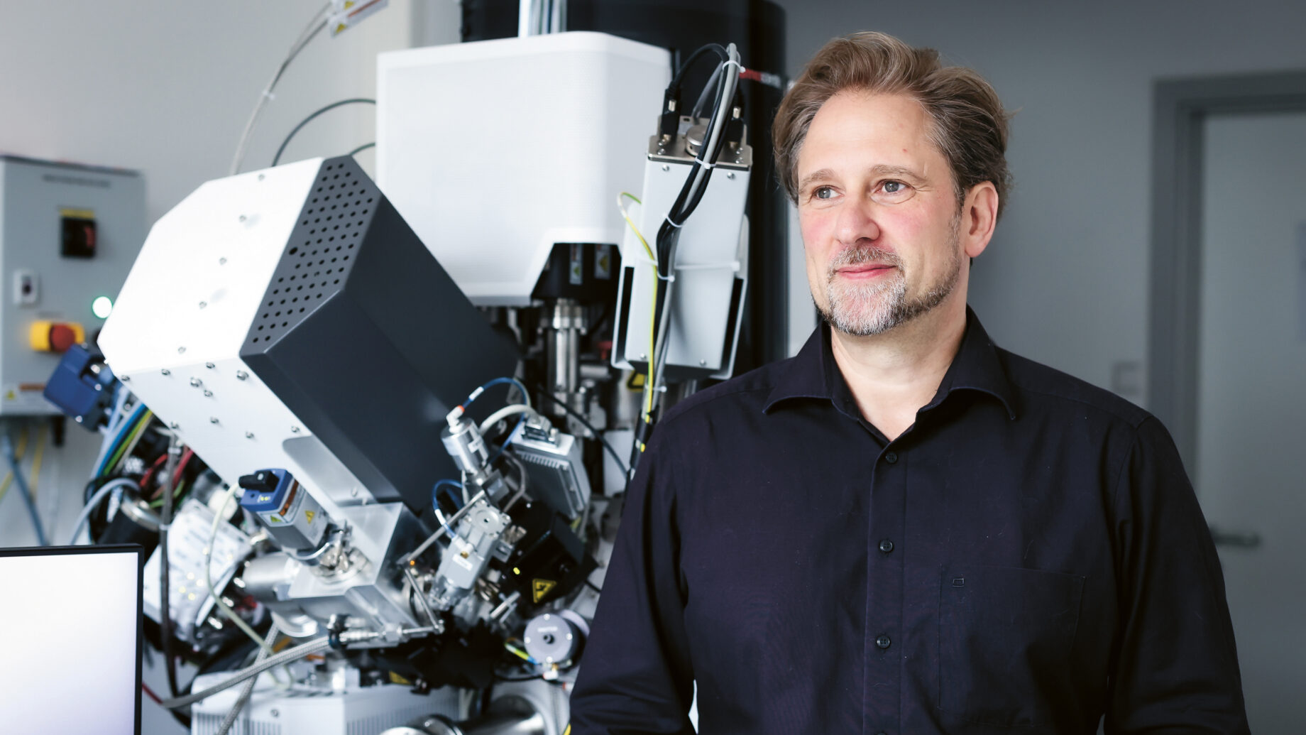 Ein Mann mit hellbraunem Haar und Bart steht in einem Labor vor hochmodernen wissenschaftlichen Geräten, trägt ein schwarzes Hemd und blickt leicht nach rechts, um nachdenklich zu wirken.