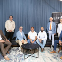 Neun Männer posieren für ein Gruppenfoto in einem modernen Raum mit blauen Vorhängen, einem runden schwarzen Couchtisch und einem "SPIRIT"-Banner. Vier stehen, fünf sitzen, alle in Business- oder eleganter Freizeitkleidung gekleidet.