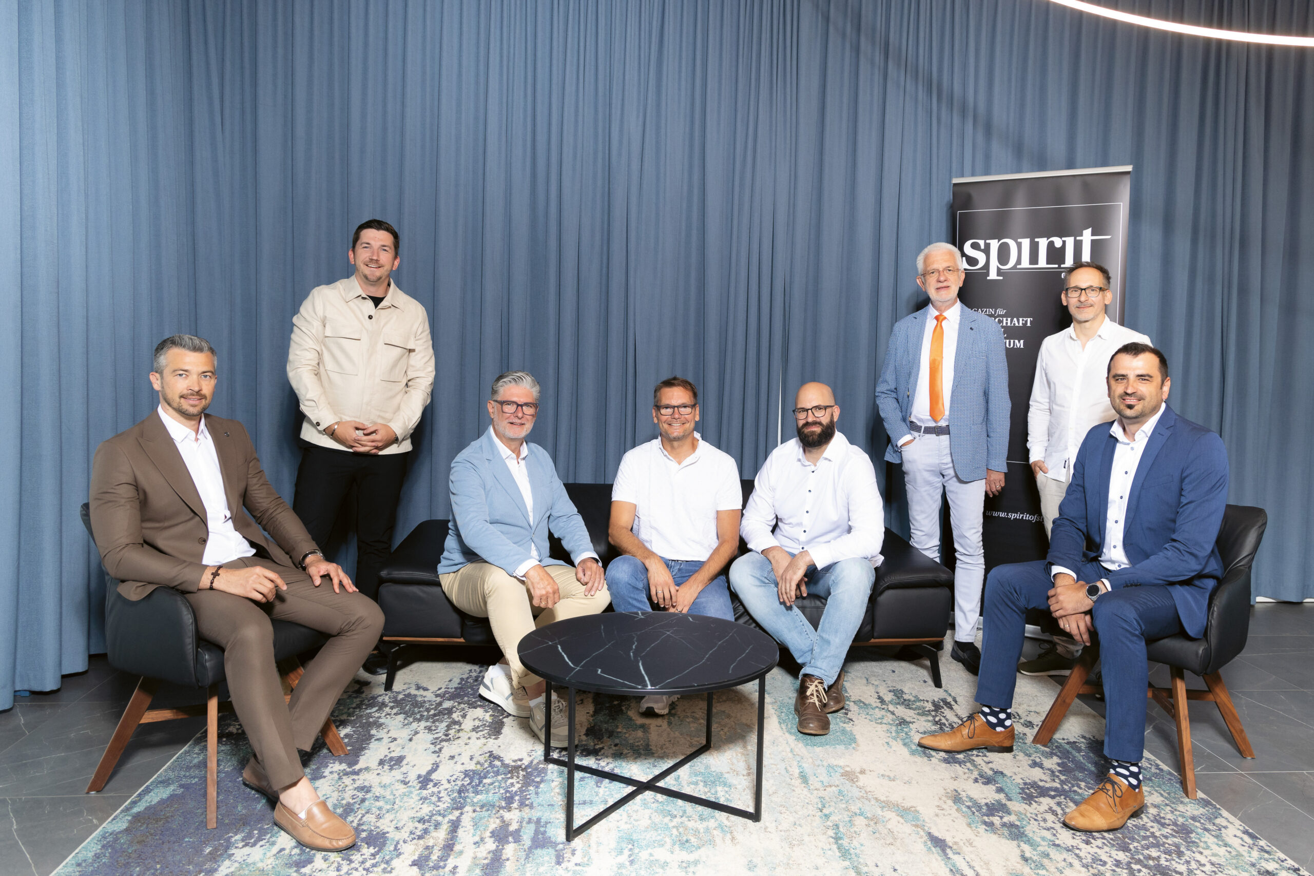 Neun Männer posieren für ein Gruppenfoto in einem modernen Raum mit blauen Vorhängen, einem runden schwarzen Couchtisch und einem "SPIRIT"-Banner. Vier stehen, fünf sitzen, alle in Business- oder eleganter Freizeitkleidung gekleidet.