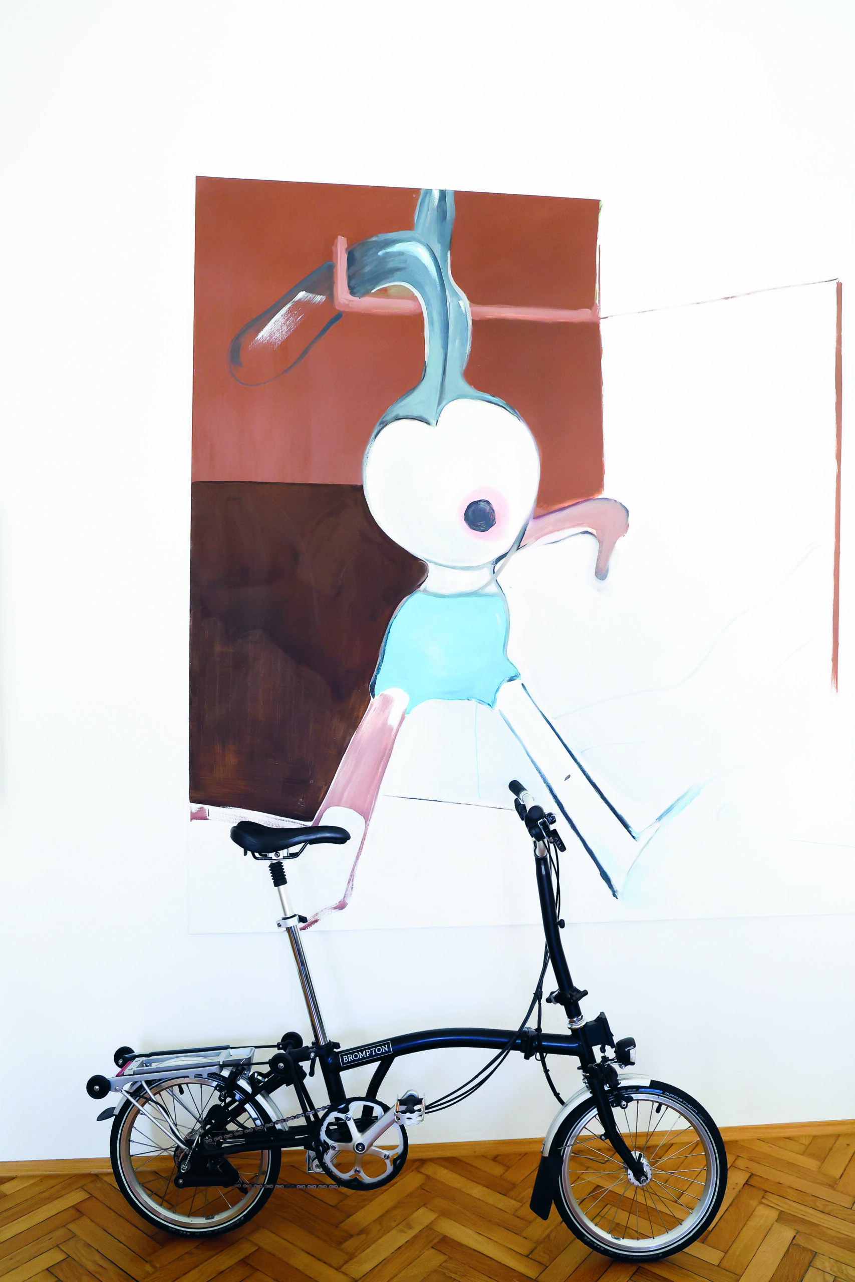 Ein schwarzes Klapprad ist vor einem großen abstrakten Gemälde geparkt. Das Gemälde zeigt eine Figur mit einem zwiebelförmigen Kopf, blauen Shorts und ausgestreckten Armen auf weißem und braunem Hintergrund.