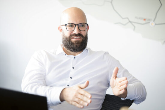 Ein bärtiger Mann mit Brille, der ein weißes Hemd trägt, gestikuliert mit seinen Händen, während er spricht. Er sitzt in einem Innenraum, hinter ihm ist eine verschwommene Landkarte an der Wand zu sehen.