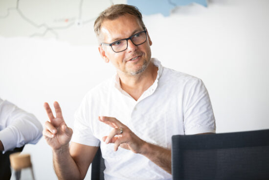Ein Mann mit Brille und weißem Hemd sitzt da und gestikuliert mit beiden Händen, wobei er mit den Fingern Anführungszeichen bildet. Er scheint in ein Gespräch vertieft zu sein. Der Hintergrund ist hell und unscharf.