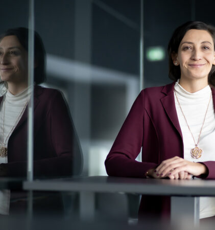 Eine Frau in einem kastanienbraunen Blazer und weißem Oberteil sitzt lächelnd mit gefalteten Händen an einem Tisch. Ihr Spiegelbild erscheint auf einer Glaswand neben ihr in einem modernen, schwach beleuchteten Raum.