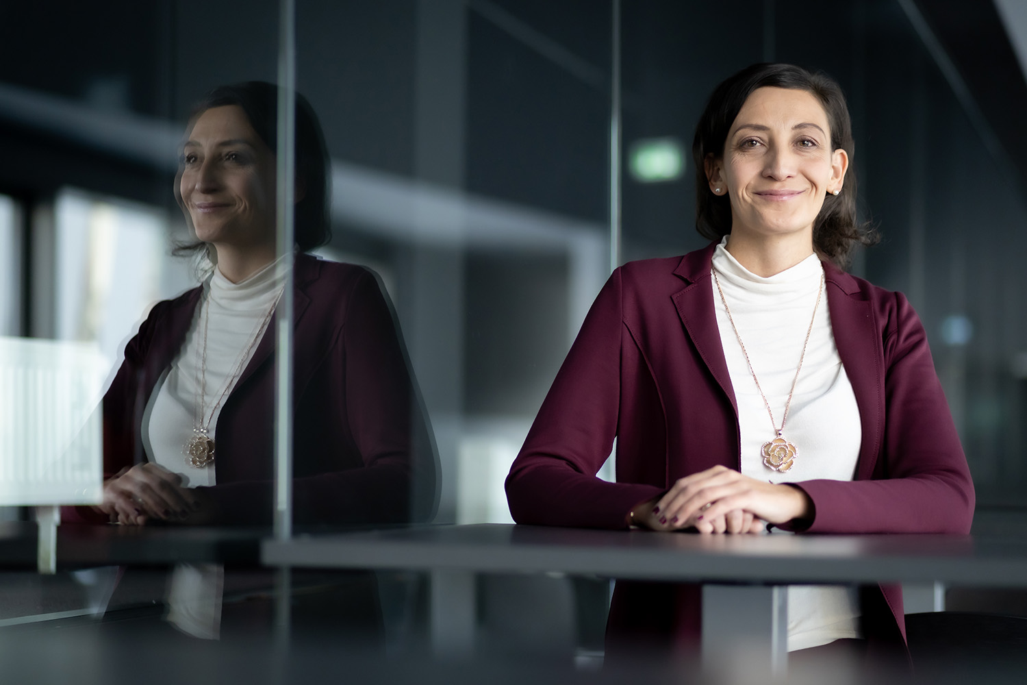 Eine Frau in einem kastanienbraunen Blazer und weißem Oberteil sitzt lächelnd mit gefalteten Händen an einem Tisch. Ihr Spiegelbild erscheint auf einer Glaswand neben ihr in einem modernen, schwach beleuchteten Raum.