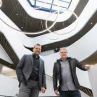 Zwei Männer in legerer Geschäftskleidung stehen in einem Innenraum unter modernen, kreisrunden Beleuchtungskörpern, vor einem architektonischen Hintergrund mit geschwungenen Linien, Balkonen und einem großen Oberlicht darüber.