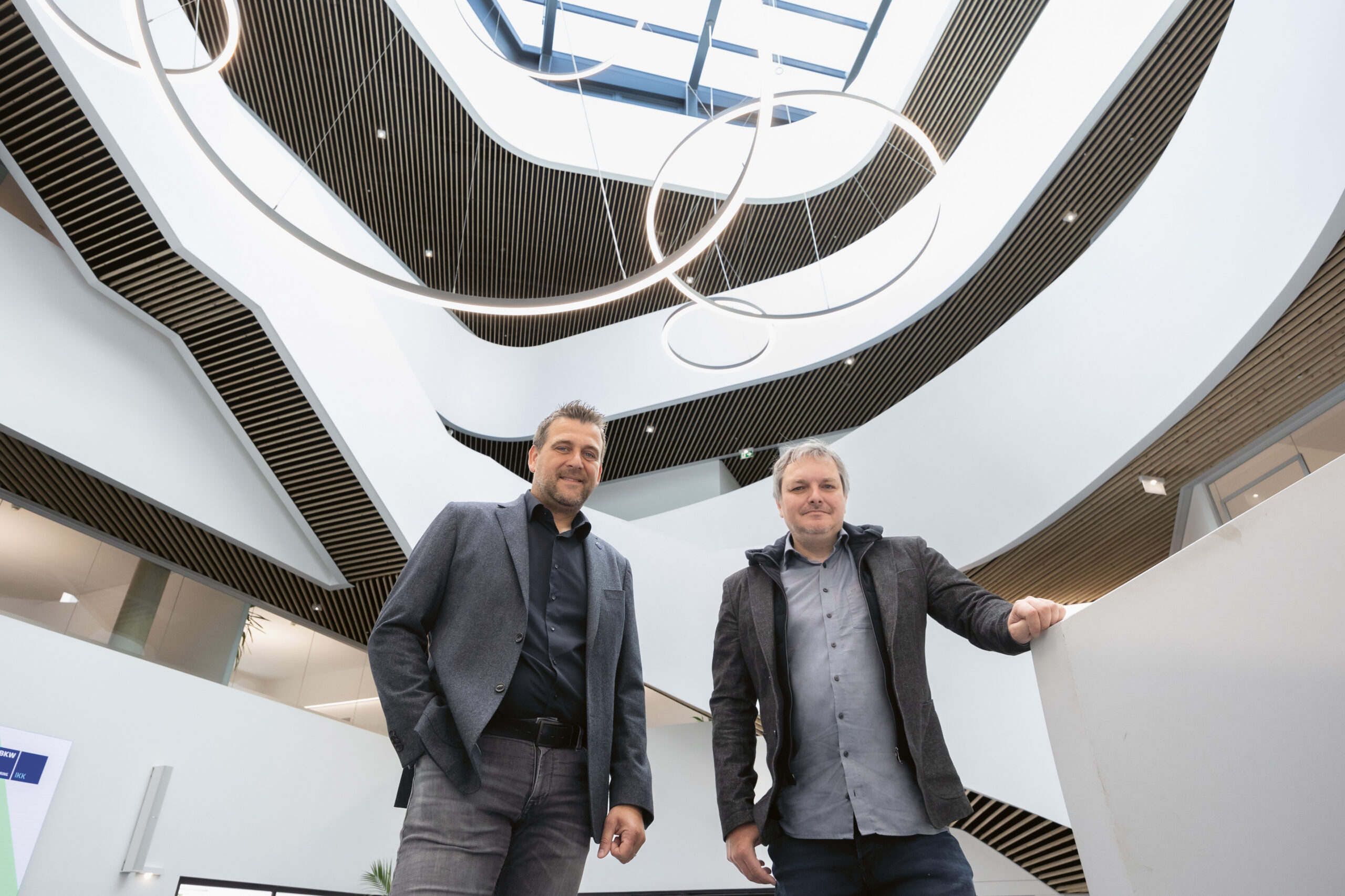 Zwei Männer in legerer Geschäftskleidung stehen in einem Innenraum unter modernen, kreisrunden Beleuchtungskörpern, vor einem architektonischen Hintergrund mit geschwungenen Linien, Balkonen und einem großen Oberlicht darüber.