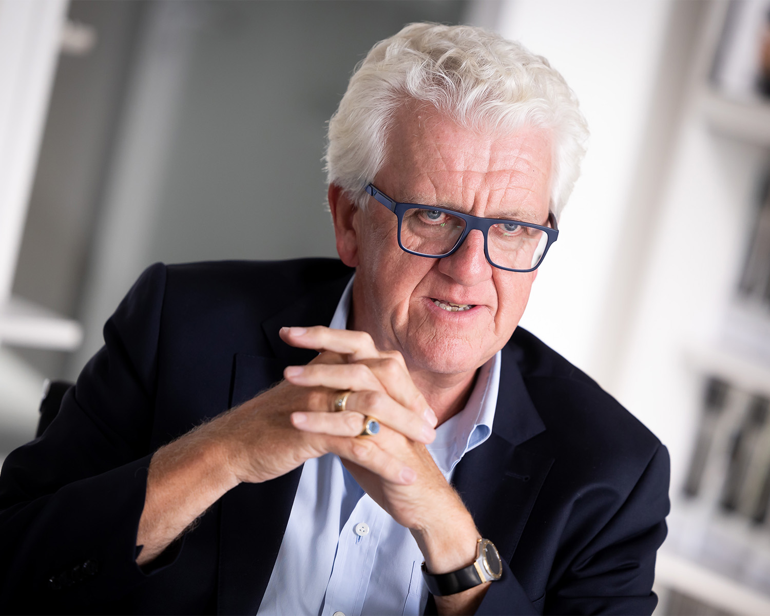 Ein älterer Mann mit weißem Haar und Brille sitzt in einem dunklen Anzug und einem hellblauen Hemd im Haus. Er blickt leicht zur Seite, hat die Hände gefaltet und wirkt nachdenklich. Der Hintergrund ist leicht unscharf.