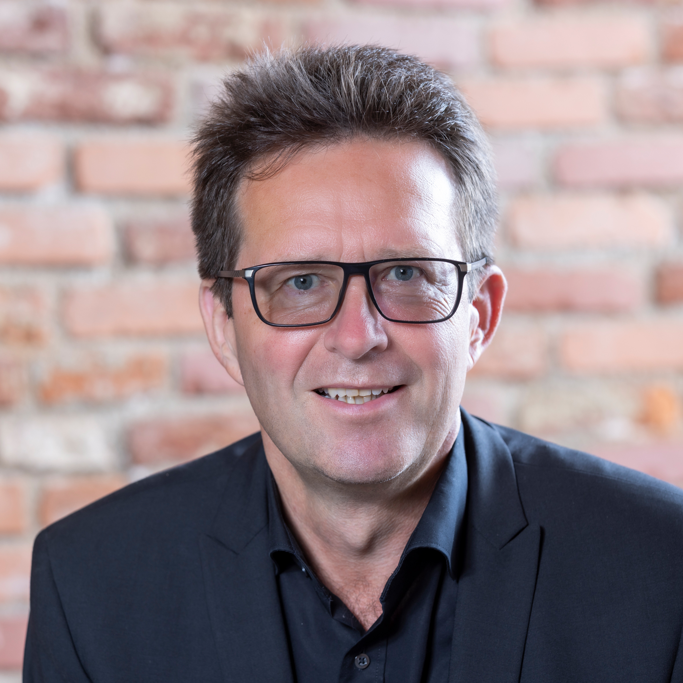 Ein Mann mit kurzen braunen Haaren und Brille, der einen dunklen Anzug und ein schwarzes Hemd trägt, lächelt in die Kamera. Der Hintergrund ist eine unscharfe Backsteinmauer.
