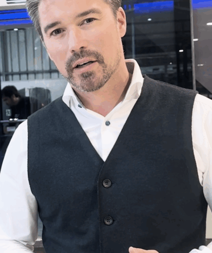 Ein Mann mit kurzem, dunklem Haar und gestutztem Bart trägt ein weißes Hemd und eine dunkle Weste. Er steht in einem Innenraum und blickt in die Kamera, wobei er mit einer Hand eine Geste ausführt. Im Hintergrund sind Glasscheiben und eine arbeitende Person zu sehen.