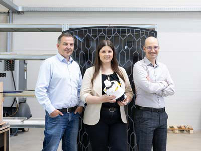 Drei Personen stehen in einem Innenraum vor einer Industrieanlage. Die Frau in der Mitte hält ein weißes 3D-gedrucktes Objekt, während die beiden Männer zu beiden Seiten in entspannter Haltung stehen und in die Kamera lächeln.