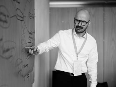 Ein Mann mit Brille und weißem Hemd zeigt während einer Präsentation oder Besprechung auf ein Diagramm auf einem Flipchart. Er trägt ein Schlüsselband um seinen Hals und scheint etwas zu erklären.