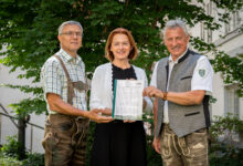 Drei lächelnde Erwachsene stehen im Freien vor einem Baum und halten gemeinsam ein Dokument. Die beiden Männer tragen traditionelle deutsche oder österreichische Trachten, die Frau in der Mitte trägt ein schwarz-weißes Outfit.