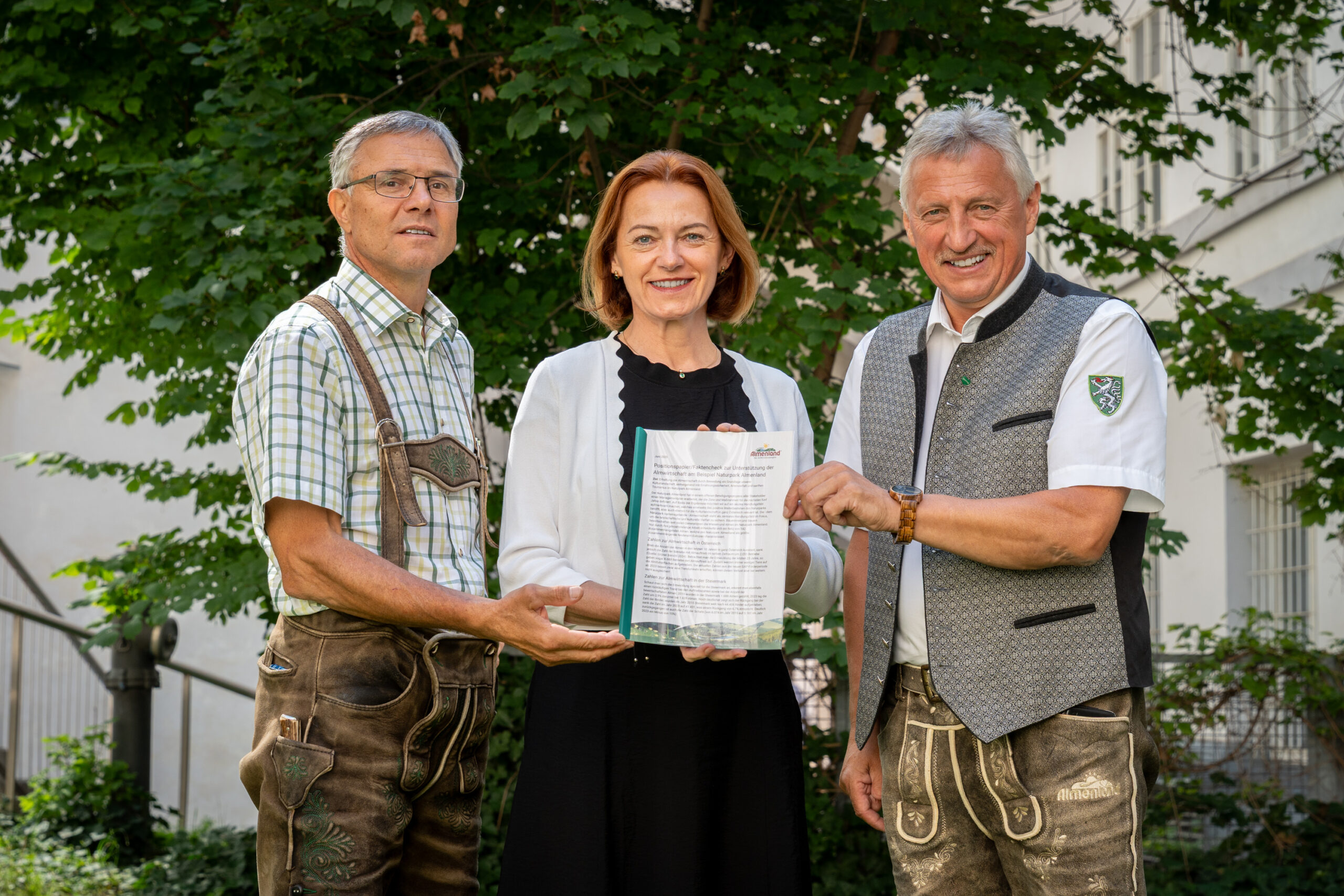 Drei lächelnde Erwachsene stehen im Freien vor einem Baum und halten gemeinsam ein Dokument. Die beiden Männer tragen traditionelle deutsche oder österreichische Trachten, die Frau in der Mitte trägt ein schwarz-weißes Outfit.