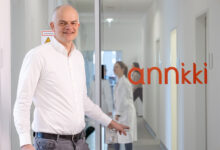 Ein Mann in einem weißen Hemd steht an einer Glastür mit der Aufschrift "annkki" und lächelt in die Kamera. Drinnen arbeiten Menschen in Laborkitteln in einem hellen Flur, der den Fokus des Unternehmens auf Nachhaltigkeit in Immobilien widerspiegelt.