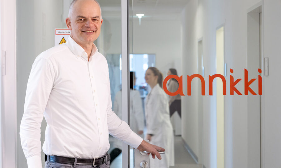 Ein Mann in einem weißen Hemd steht an einer Glastür mit der Aufschrift "annkki" und lächelt in die Kamera. Drinnen arbeiten Menschen in Laborkitteln in einem hellen Flur, der den Fokus des Unternehmens auf Nachhaltigkeit in Immobilien widerspiegelt.