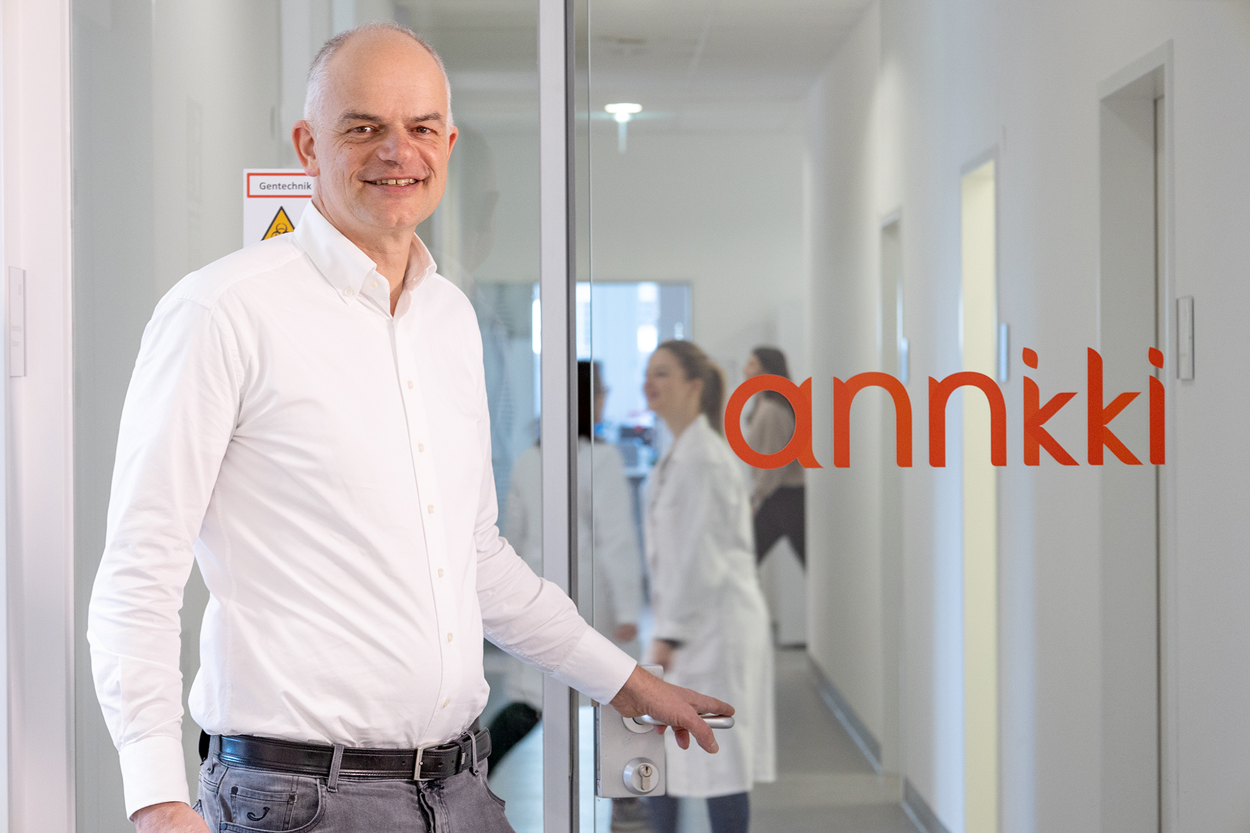 Ein Mann in einem weißen Hemd steht an einer Glastür mit der Aufschrift "annkki" und lächelt in die Kamera. Drinnen arbeiten Menschen in Laborkitteln in einem hellen Flur, der den Fokus des Unternehmens auf Nachhaltigkeit in Immobilien widerspiegelt.