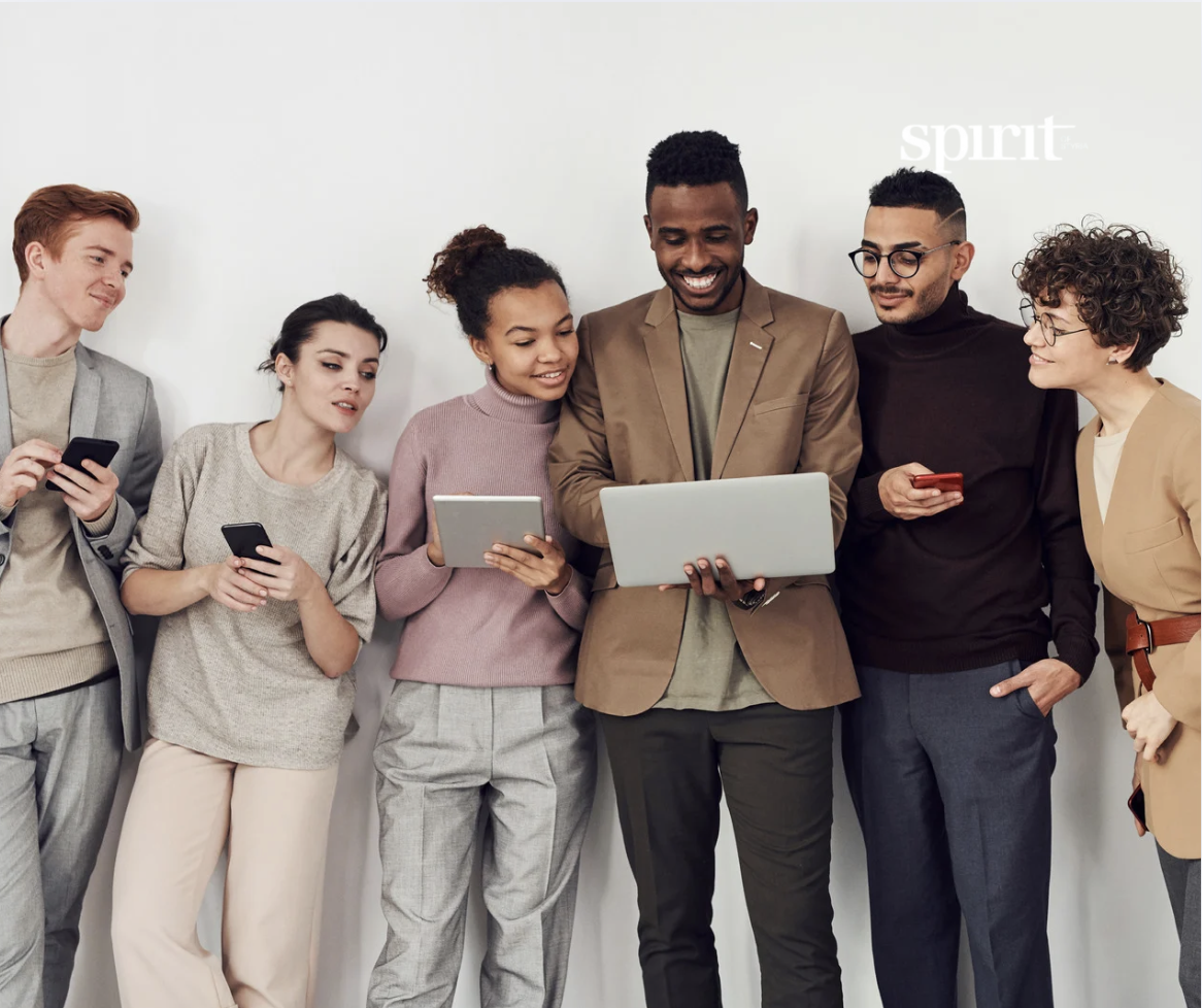 Eine bunt gemischte Gruppe von sechs Personen steht lächelnd zusammen und schaut auf einen Laptop. Einige halten Telefone und ein Tablet in der Hand. Sie wirken kooperativ und engagiert und stehen vor einem schlichten weißen Hintergrund mit dem Wort "Spirit" in der Ecke.